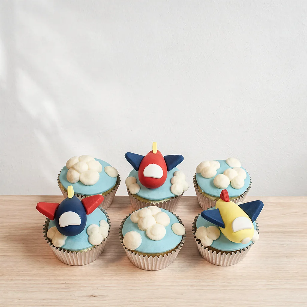 Six cupcakes with blue icing, white clouds, and three colorful fondant airplanes on top