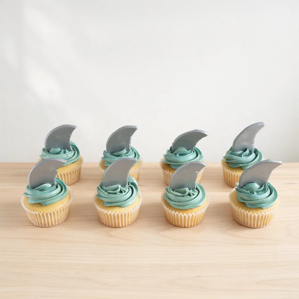 Eight vanilla cupcakes with teal frosting and edible silver shark fin toppers